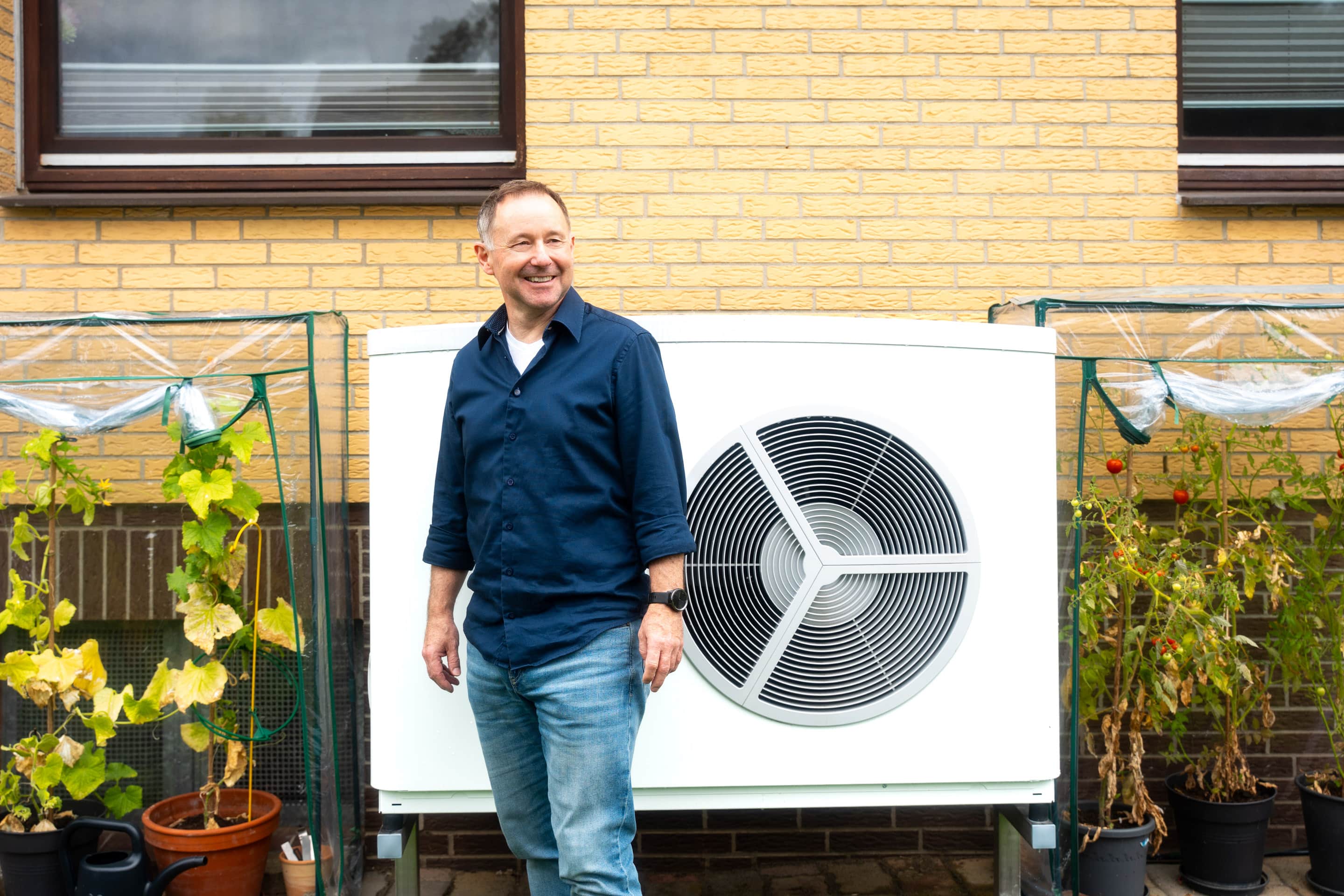 Mann steht vor einer Wärmepumpe im Garten und lächelt
